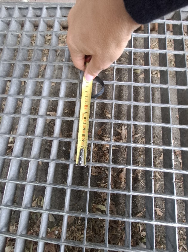 Miara sprawdza szerokość otworów metalowej kraty na dojściu do budynku.