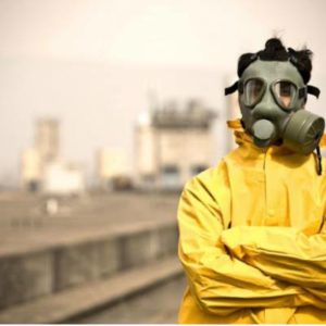 Osoba w żółtym kombinezonie ochronnym i masce przeciwgazowej na twarzy na tle słabo widocznej fabryki