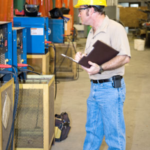 Na zdjęciu widać mężczyznę w żółtym kasku na głowie sprawdzającego maszynę w fabryce. Mężczyzna trzyma w rękach podkładkę z papierem i notuje uwagi powstałe podczas kontroli.